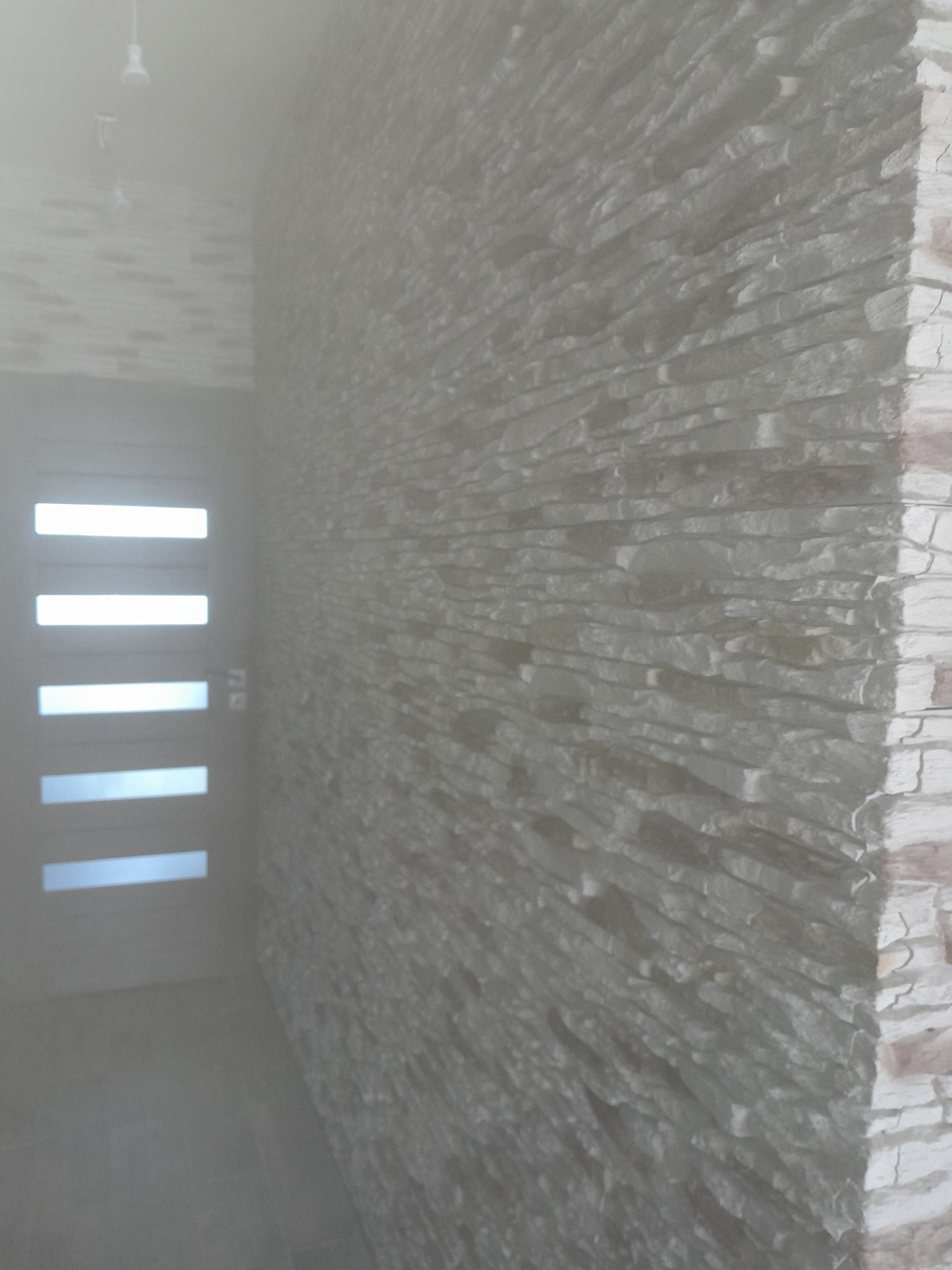 Ściana wykończona szarym kamieniem dekoracyjnym, widoczna z boku, z fragmentem drzwi z poziomymi przeszkleniami w tle.