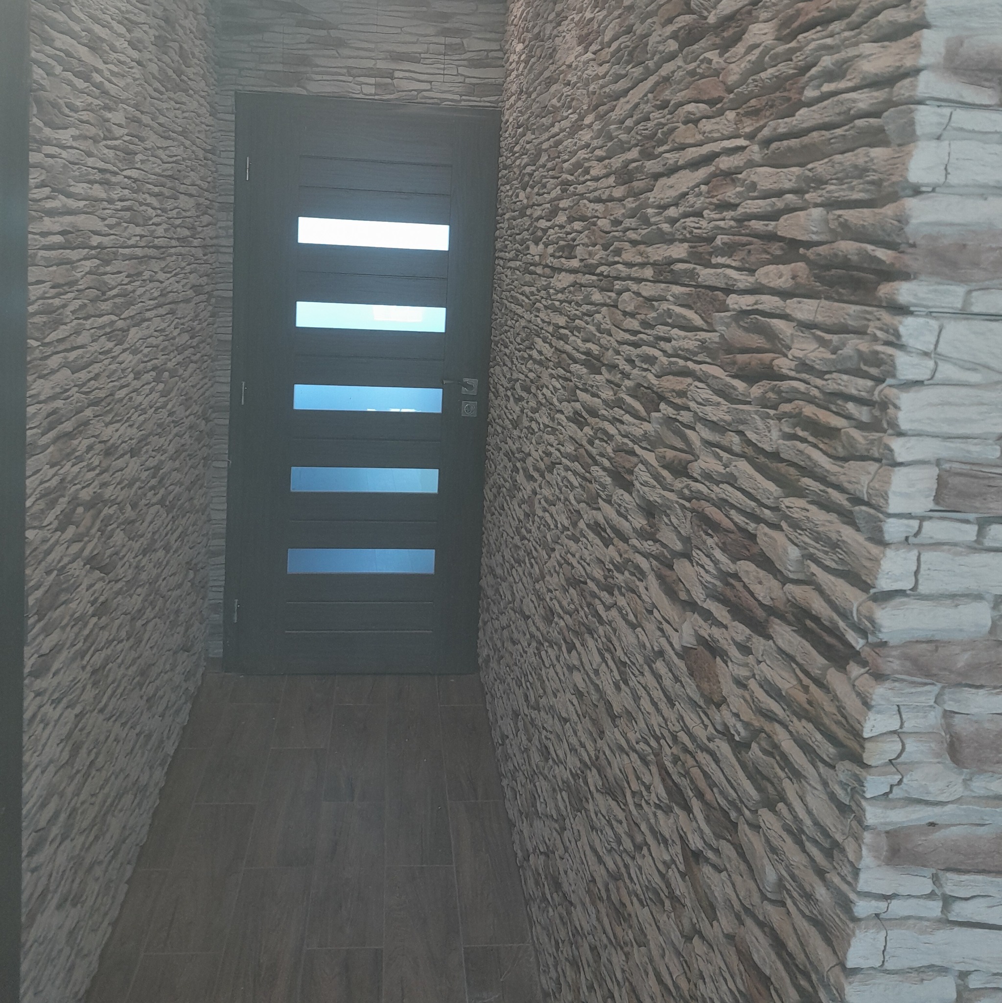 Zamontowane drzwi wewnętrzne z przeszkleniami w Koło, otoczone dekoracyjną ścianą z kamienia imitującego naturalny łupek, dopasowaną kolorystycznie do podłogi z płytek ceramicznych.