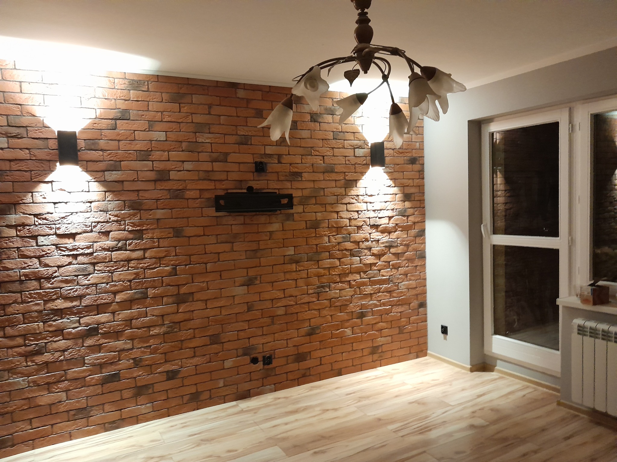 Salon z ceglaną ścianą, oświetleniem punktowym i drewnianą podłogą, widok na okno z grzejnikiem.