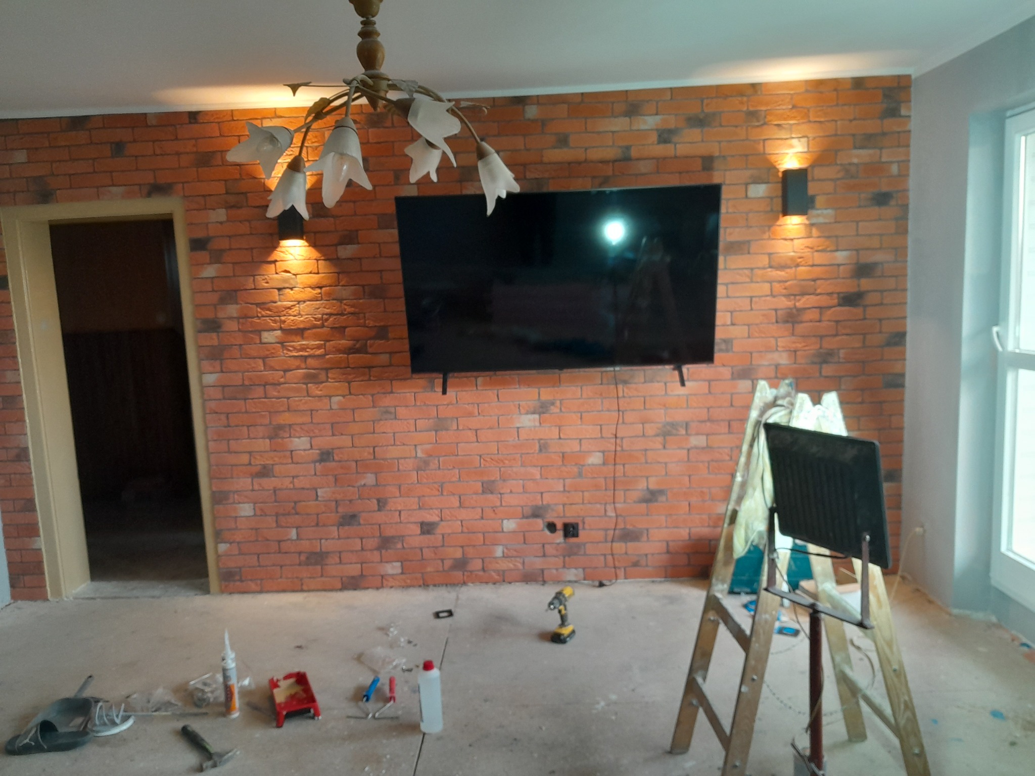 Pokój w trakcie prac wykończeniowych: ściana z cegły z zamontowanym telewizorem, wiszące lampy, drabina, narzędzia, surowa podłoga.