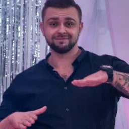 Uśmiechnięty mężczyzna z tatuażem na ramieniu, w czarnej koszuli, prezentuje gestem dłoni przestrzeń, tło z srebrnymi frędzlami i białym materiałem.