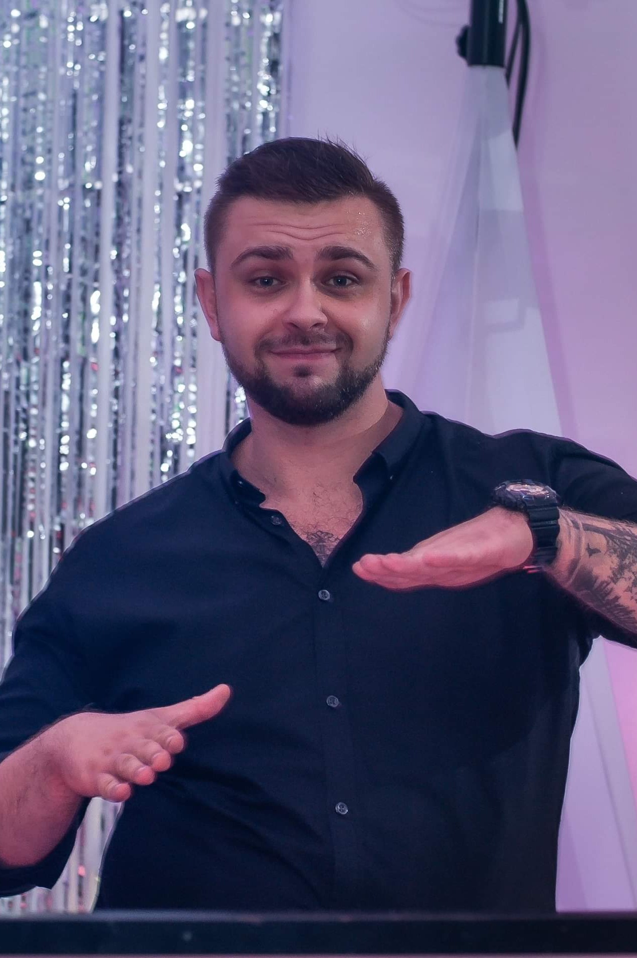 Uśmiechnięty mężczyzna z tatuażem na ramieniu, w czarnej koszuli, prezentuje gestem dłoni przestrzeń, tło z srebrnymi frędzlami i białym materiałem.