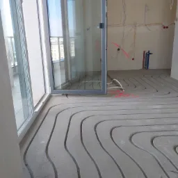 Wylewka betonowa z wyfrezowanymi rowkami pod instalację ogrzewania podłogowego, widoczne rury instalacji elektrycznej i wodnej na ścianie w surowym stanie.