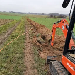 Pomarańczowa mini koparka Kubota KX019 w trakcie wykonywania wykopu w ziemi na tle pola i pochmurnego nieba; widoczny długi, wąski wykop z ułożonym wzdłuż niego pomarańczowym przewodem.