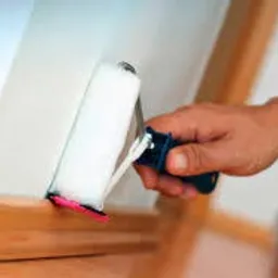 Malowanie narożnika ściany białym wałkiem z fioletową podkładką, widoczna dłoń i fragment drewnianej listwy przypodłogowej.