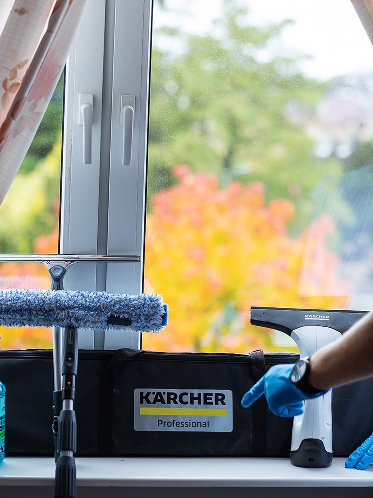 Profesjonalne narzędzia Kärcher, w tym myjka i ściągaczka, na parapecie przed brudną szybą okienną z jesiennym krajobrazem. Ręka w niebieskiej rękawiczce wskazuje na torbę Kärcher Professional.