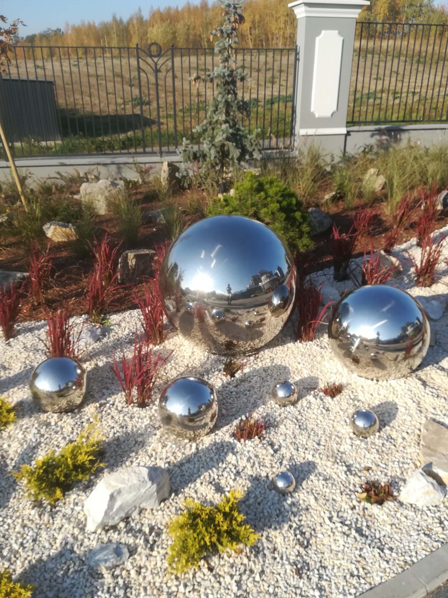 Ogród w Kłodzku z dekoracyjnymi, lustrzanymi kulami różnej wielkości, ułożonymi na białym żwirze wśród niskich roślin i kamieni, z ogrodzeniem w tle.