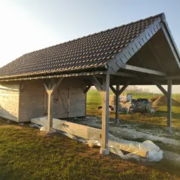 Drewniana konstrukcja z dwuspadowym dachem pokrytym ciemną dachówką, widoczna w promieniach zachodzącego słońca na tle zielonego pola, z materiałami budowlanymi zabezpieczonymi folią w pierwszym...