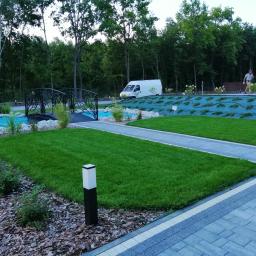 Garden Service Ogrody Adrian Kowal - Nowoczesny ogród z równo przystrzyżonym trawnikiem, ozdobnym mostkiem nad sztucznym strumieniem, wybrukowanym chodnikiem, oświetleniem ogrodowym i roślinnością ozdobną, w tle zaparkowany biały van.