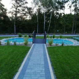 Ogród z dwoma małymi stawami połączonymi mostkiem, otoczone kamieniami i roślinnością, z wybrukowaną ścieżką prowadzącą do nich, trawnik i drzewa w tle.