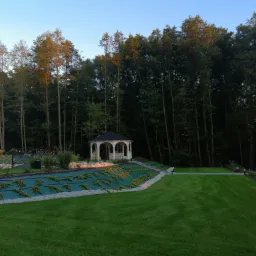 Pejzaż z altaną w ogrodzie, świeżo założony trawnik, system nawadniania pod zieloną tkaniną, oświetlenie punktowe i dekoracyjny mostek, widoczny samochód dostawczy w tle.