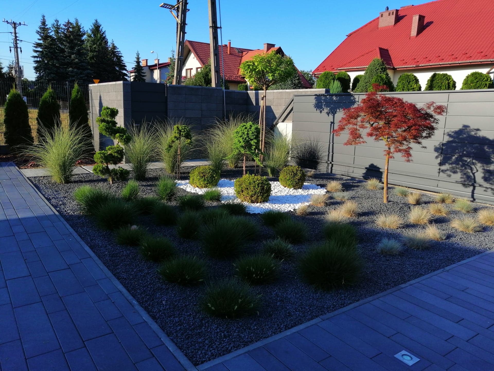 Nowoczesny ogród z geometrycznymi nasadzeniami i szarym ogrodzeniem panelowym, ozdobne trawy i drzewka na tle białego i szarego żwiru, regularny układ kostki brukowej.