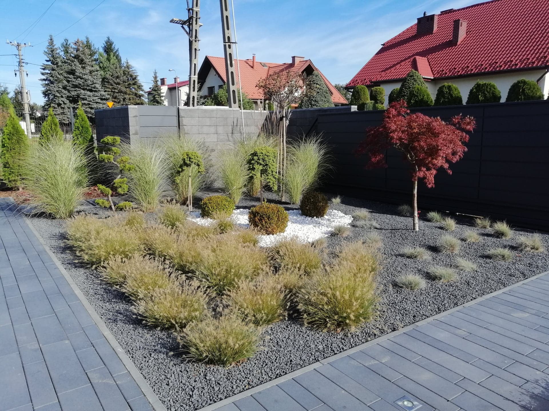 Nowoczesny ogród z ozdobnymi trawami, białymi kamieniami i czarnym żwirem, obramowany szarą kostką brukową i ciemnym ogrodzeniem panelowym, z widocznymi domami w tle.