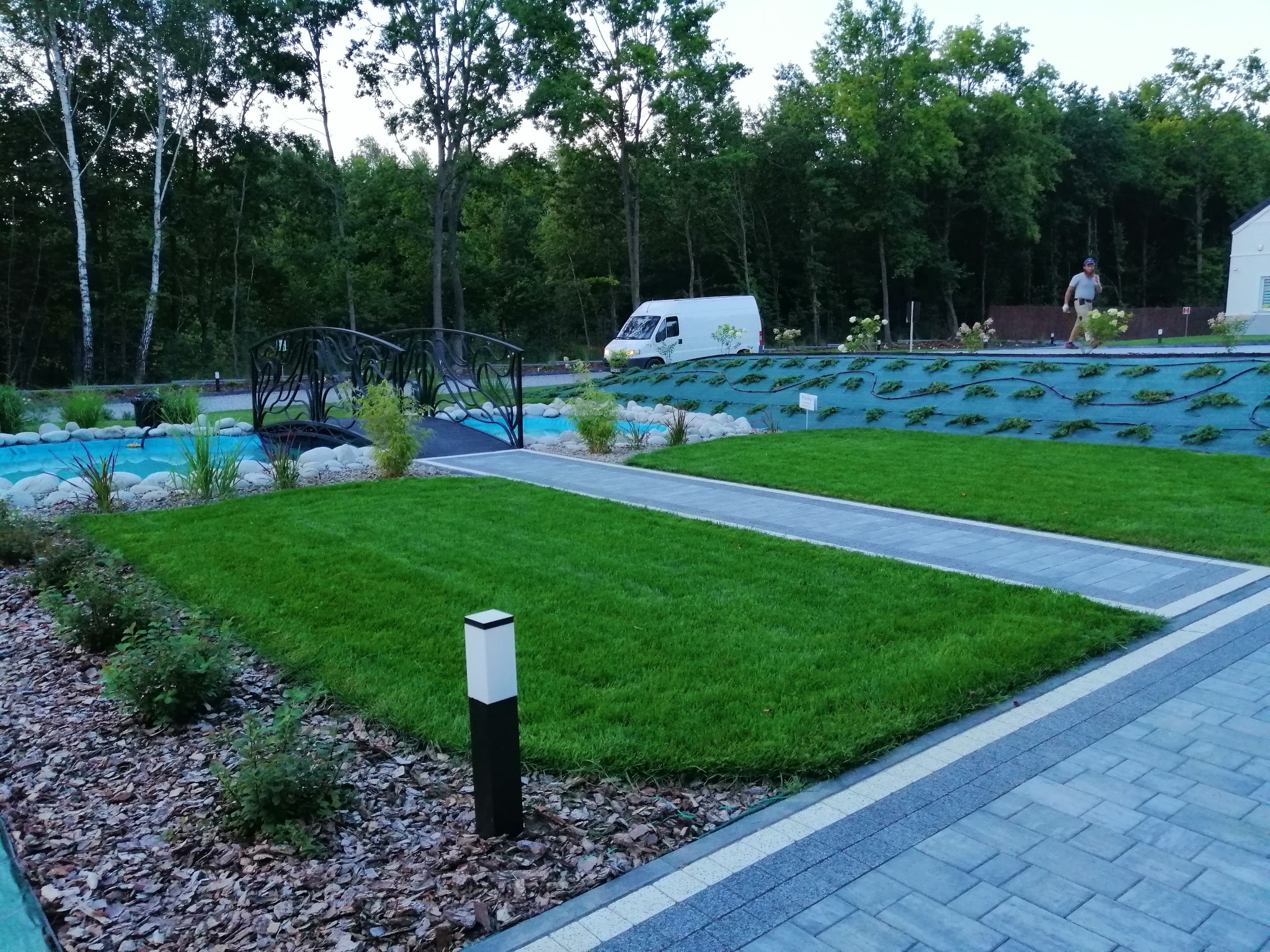 Nowoczesny ogród z równo przystrzyżonym trawnikiem, ozdobnym mostkiem nad sztucznym strumieniem, wybrukowanym chodnikiem, oświetleniem ogrodowym i roślinnością ozdobną, w tle zaparkowany biały van.