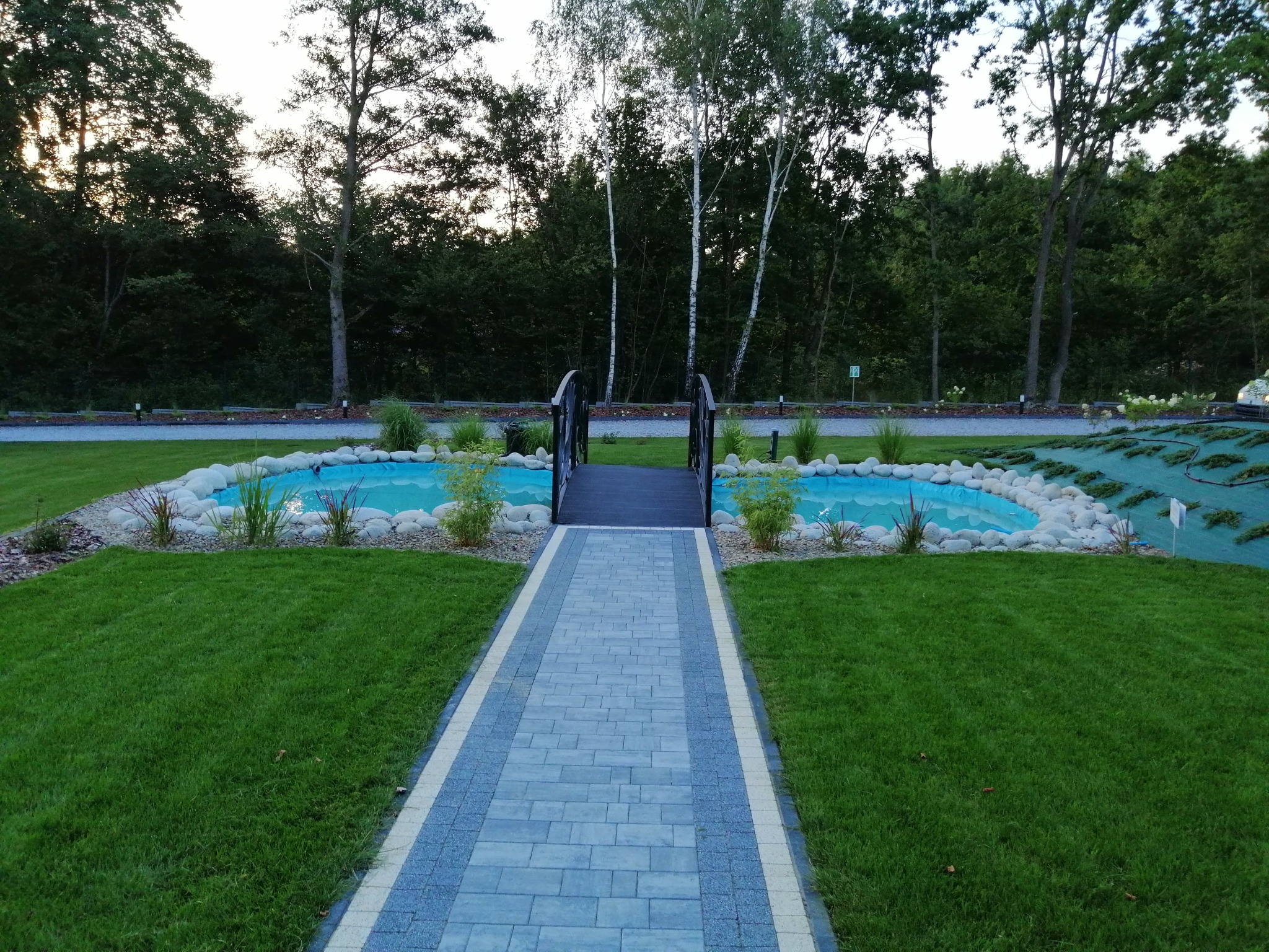 Ogród z dwoma małymi stawami połączonymi mostkiem, otoczone kamieniami i roślinnością, z wybrukowaną ścieżką prowadzącą do nich, trawnik i drzewa w tle.