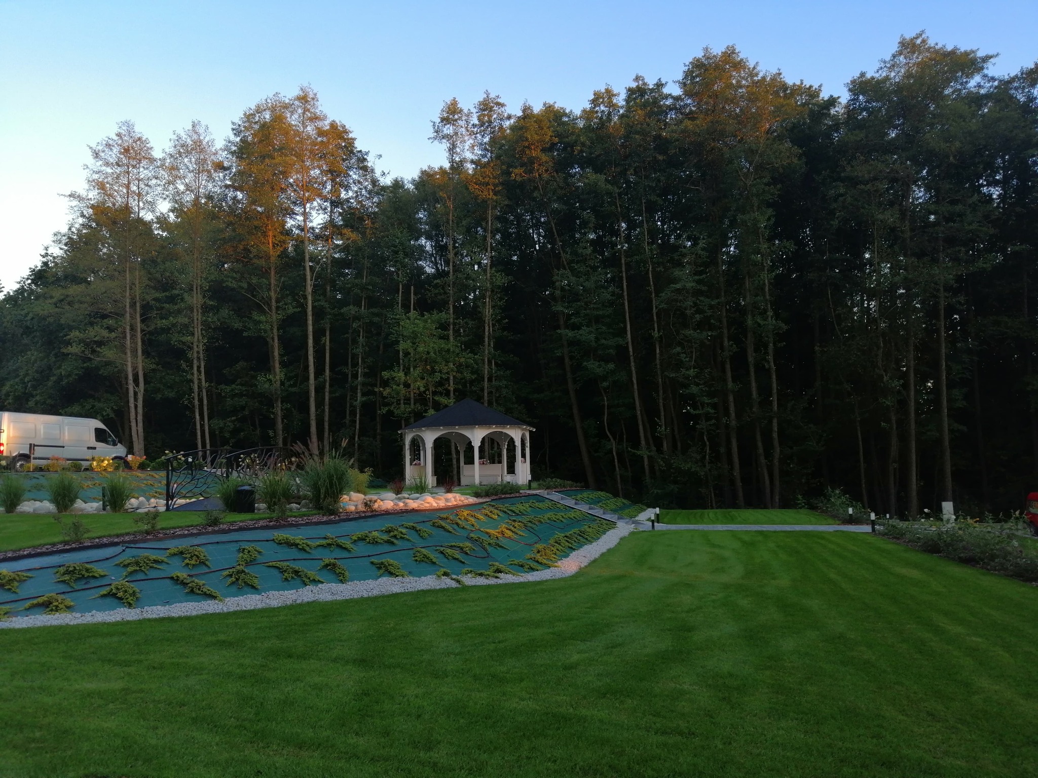 Pejzaż z altaną w ogrodzie, świeżo założony trawnik, system nawadniania pod zieloną tkaniną, oświetlenie punktowe i dekoracyjny mostek, widoczny samochód dostawczy w tle.