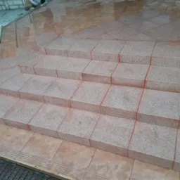 Wyłożone płytkami schody zewnętrzne z delikatnym spadkiem, z widocznymi śladami świeżego fugowania na pomarańczowo, otoczone mokrą, brązową posadzką.