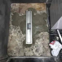 Montaż liniowego odpływu prysznicowego w surowej posadzce łazienki, widoczne ślady cementu i nier&oacute;wności podłoża, fragment buta w kadrze.