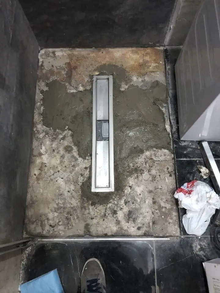 Montaż liniowego odpływu prysznicowego w surowej posadzce łazienki, widoczne ślady cementu i nierówności podłoża, fragment buta w kadrze.