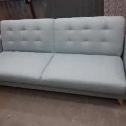 Jasnoniebieska sofa z guzikami na oparciu, ustawiona na tle blachy falistej i wzorzystej tapety, widoczne drewniane nóżki.