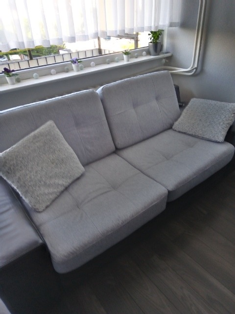 Jasnoszara, modułowa sofa z poduszkami w pokoju z drewnianą podłogą i oknem z białymi zasłonami.