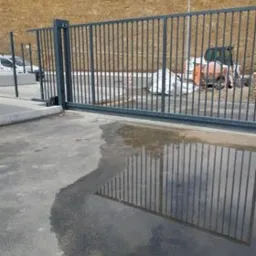 Szara, przesuwna brama wjazdowa z pionowych prętów, widoczna w odbiciu na mokrym betonowym podłożu. W tle wzgórze z zielenią i częściowo widoczne pojazdy budowlane.