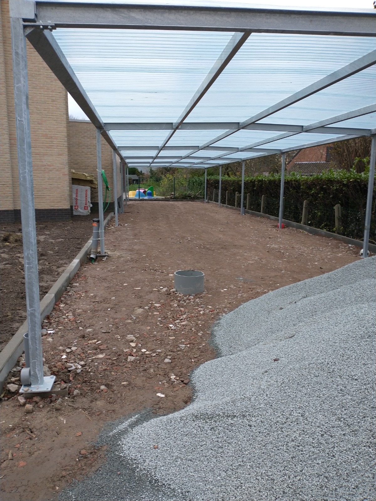 Konstrukcja zadaszenia z przezroczystego tworzywa na metalowym stelażu w trakcie budowy. Widoczne przygotowane podłoże, krawężniki i stos kruszywa.