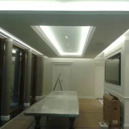 Nowoczesny sufit podwieszany z oświetleniem LED w kształcie prostokąta, z trzema punktowymi lampami, widok na przestrzeń z oknami, zabezpieczonym folią stołem i drabiną opartą o drzwi.