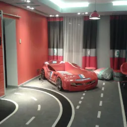 Pokój dziecięcy w motywie wyścigowym: łóżko w kształcie czerwonego samochodu wyścigowego stoi na dywanie imitującym tor, ściany pomalowane na czerwono, zasłony z motywem numeru jeden, czerwona...