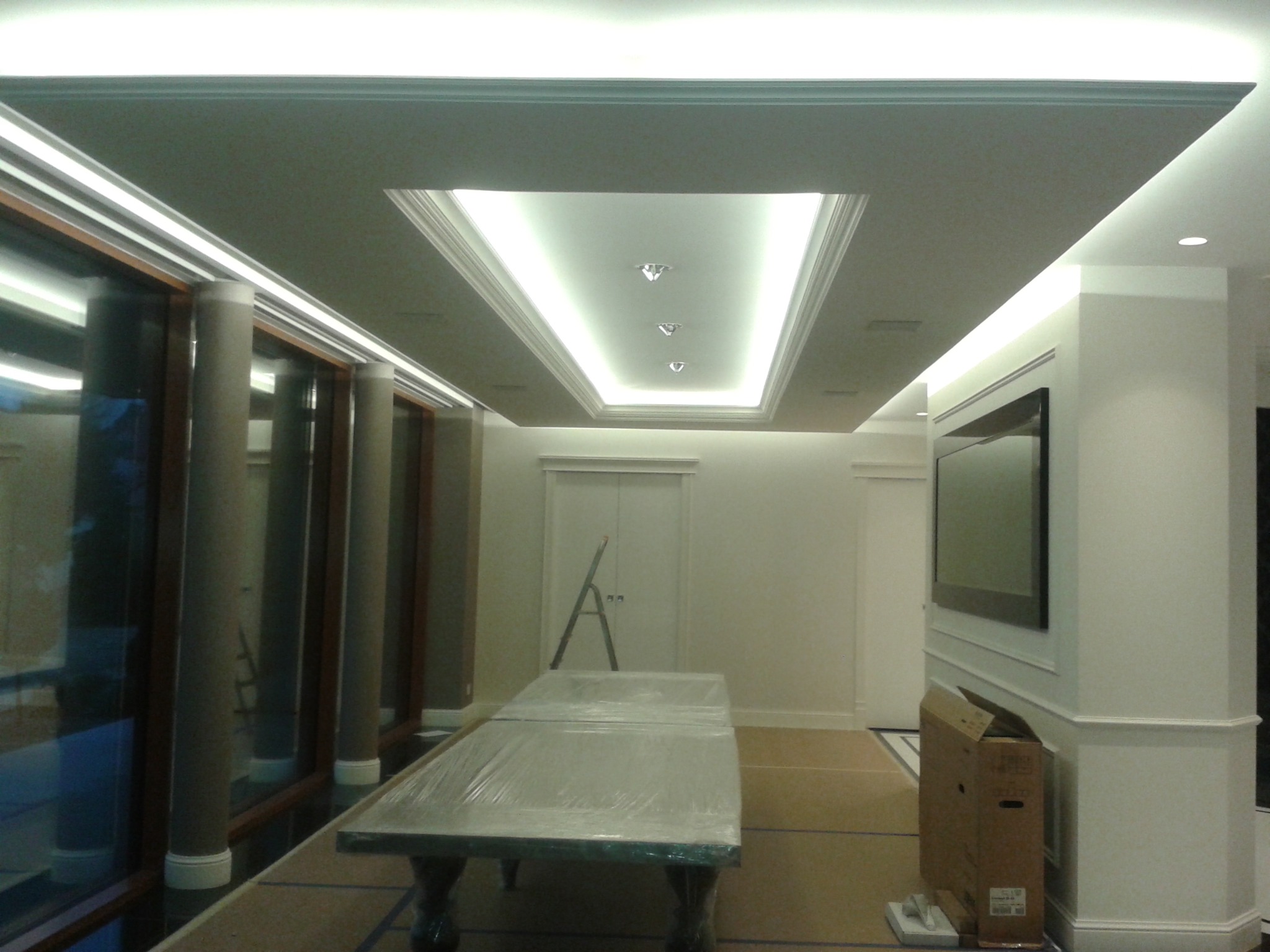 Nowoczesny sufit podwieszany z oświetleniem LED w kształcie prostokąta, z trzema punktowymi lampami, widok na przestrzeń z oknami, zabezpieczonym folią stołem i drabiną opartą o drzwi.