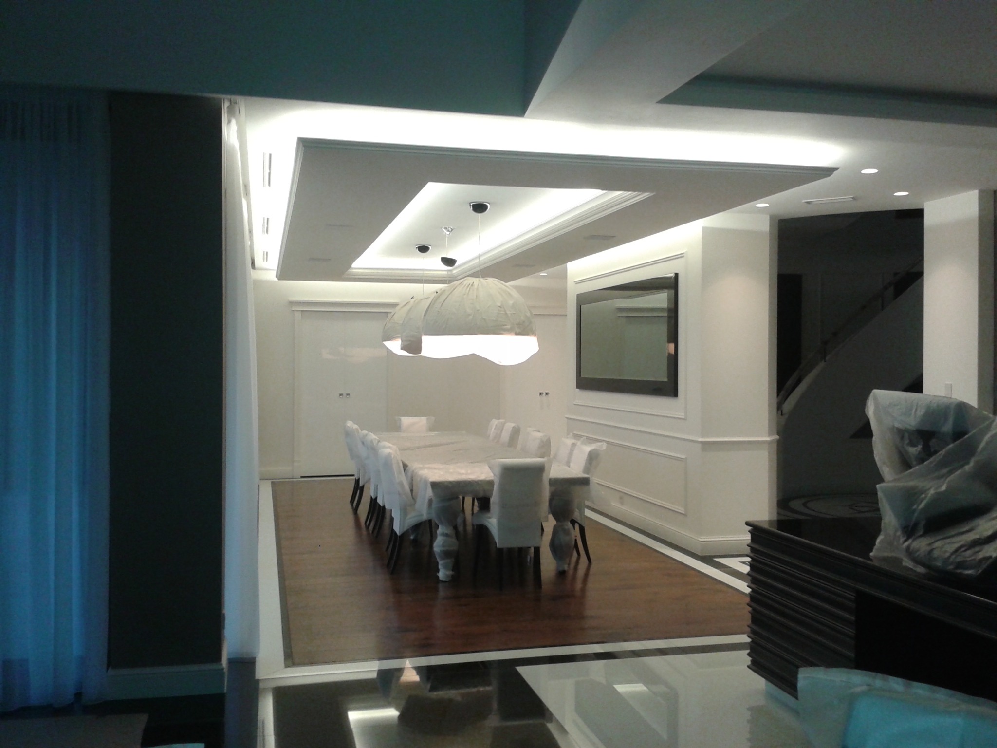 Elegancka jadalnia w stylu klasycznym z długim stołem przykrytym folią ochronną, krzesła w pokrowcach, podwieszany sufit z oświetleniem i parkiet ułożony we wzór. W tle widoczny telewizor i schody.