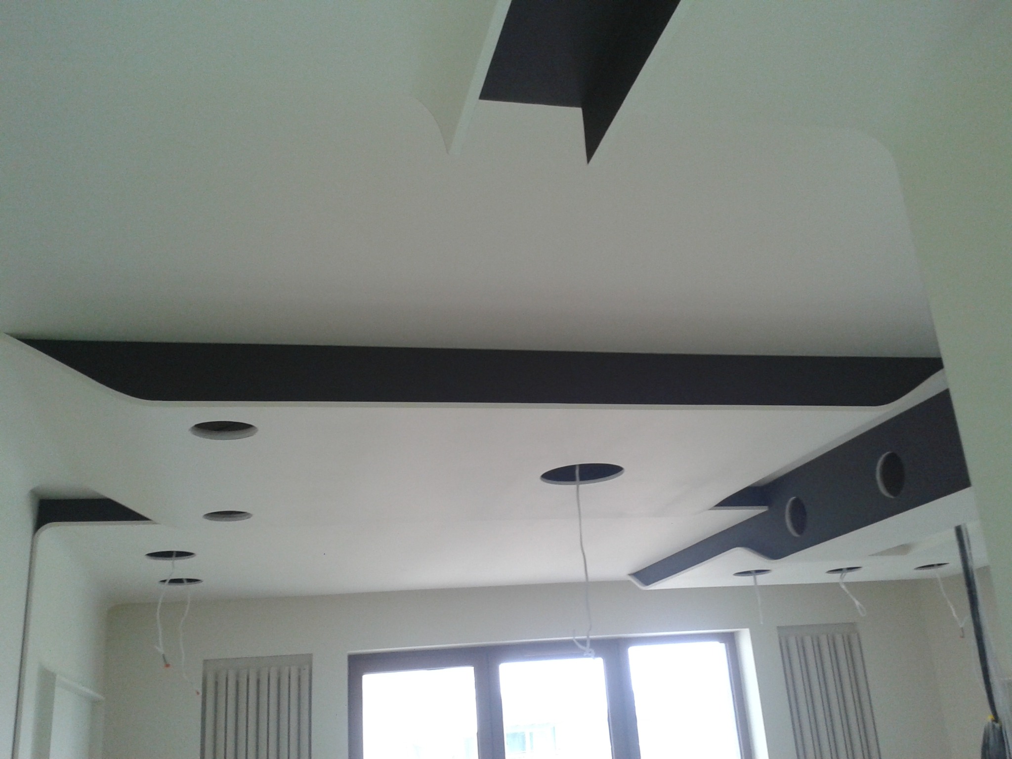 Nowoczesny, biały sufit podwieszany z czarnymi elementami dekoracyjnymi i otworami na oświetlenie, widoczne kable wystające z otworów, okno w tle.