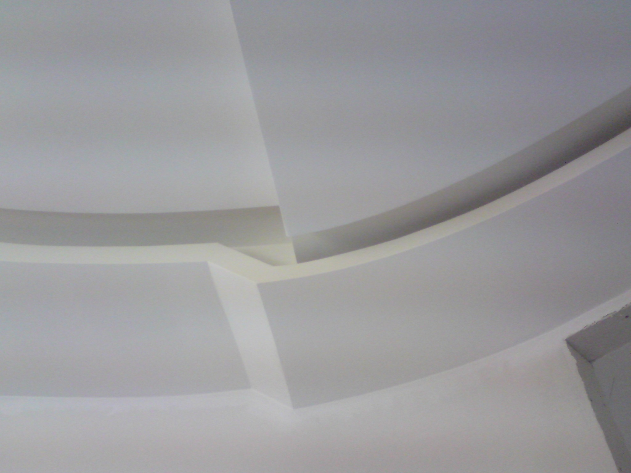 Fragment białego sufitu z dekoracyjnym, zakrzywionym elementem i widocznym fragmentem ściany w prawym dolnym rogu.
