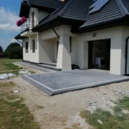 Nowoczesny taras z szarej kostki brukowej z jasną obwódką, częściowo ułożony przed garażem domu jednorodzinnego z balkonem ozdobionym różowymi kwiatami, widoczne pozostałości piasku i kostki. Przez...