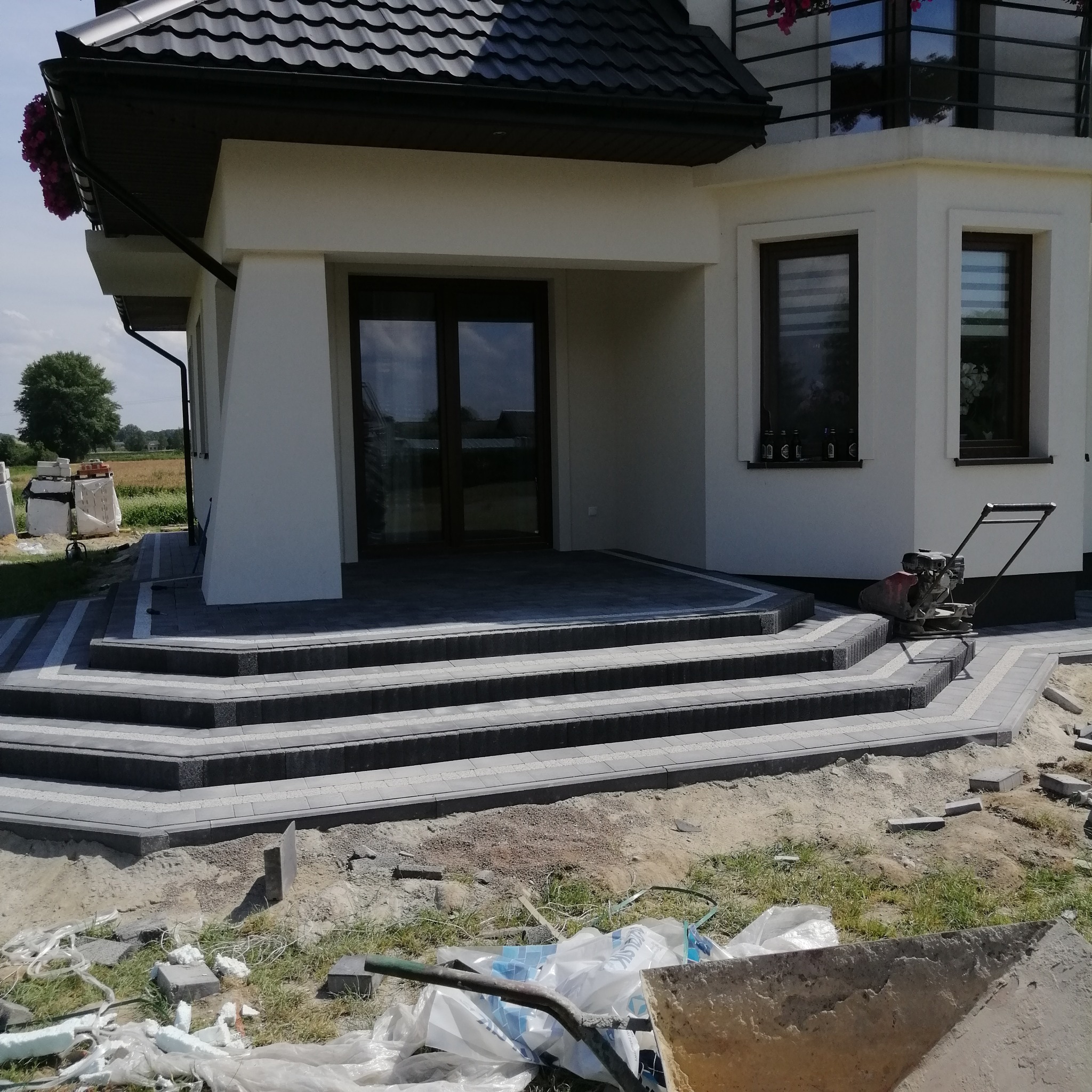 Schody wejściowe do domu w Lublinie, wyłożone kostką brukową w odcieniach szarości, z widoczną zagęszczarką i elementami placu budowy.