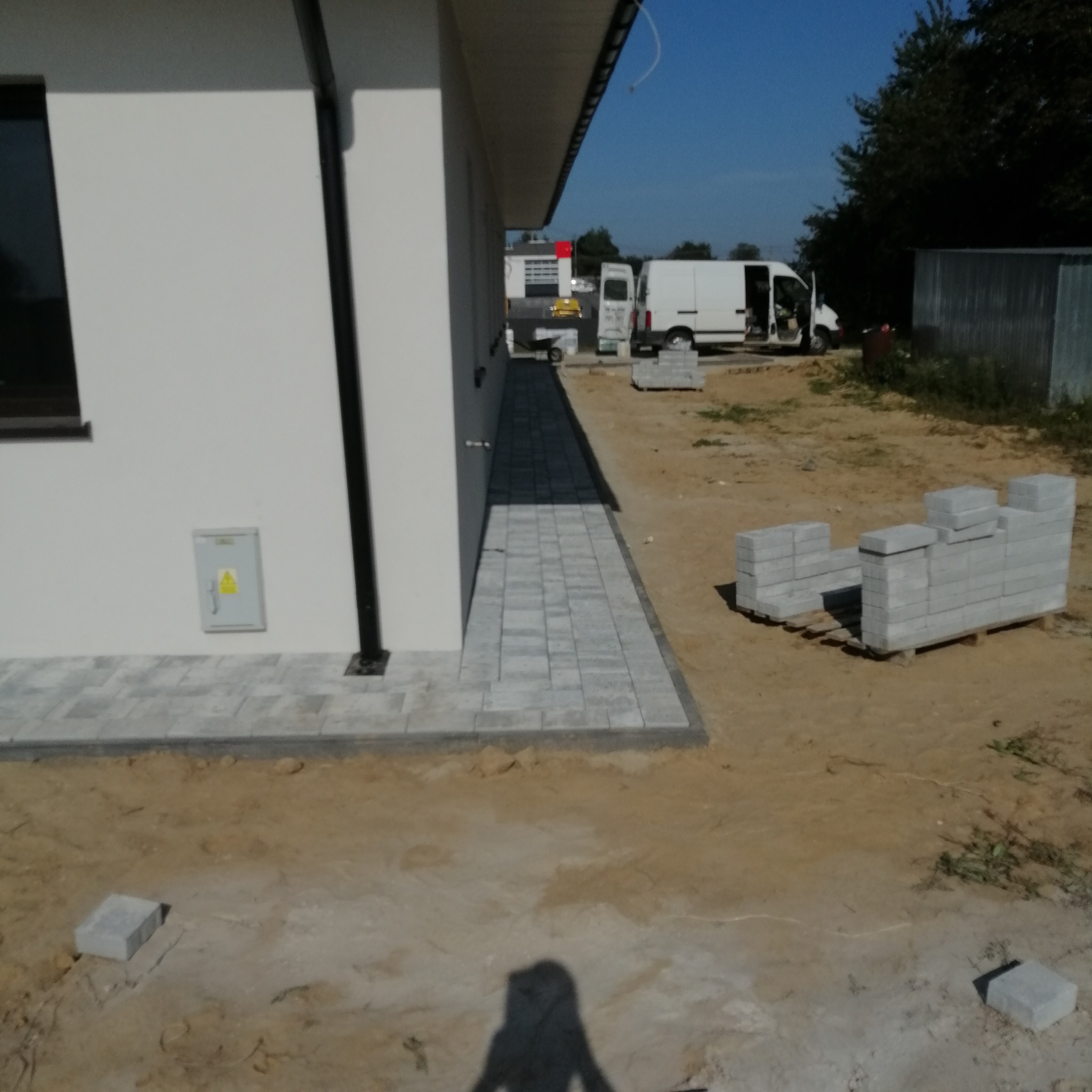 Ułożona ścieżka z szarej kostki brukowej prowadząca wzdłuż białej ściany budynku, z paletą kostki obok i samochodem dostawczym w tle.