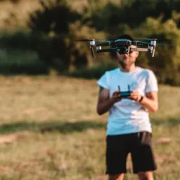 Operator drona z kontrolerem w rękach, dron Mavic 2 Pro zawieszony w powietrzu na tle łąki i drzew.