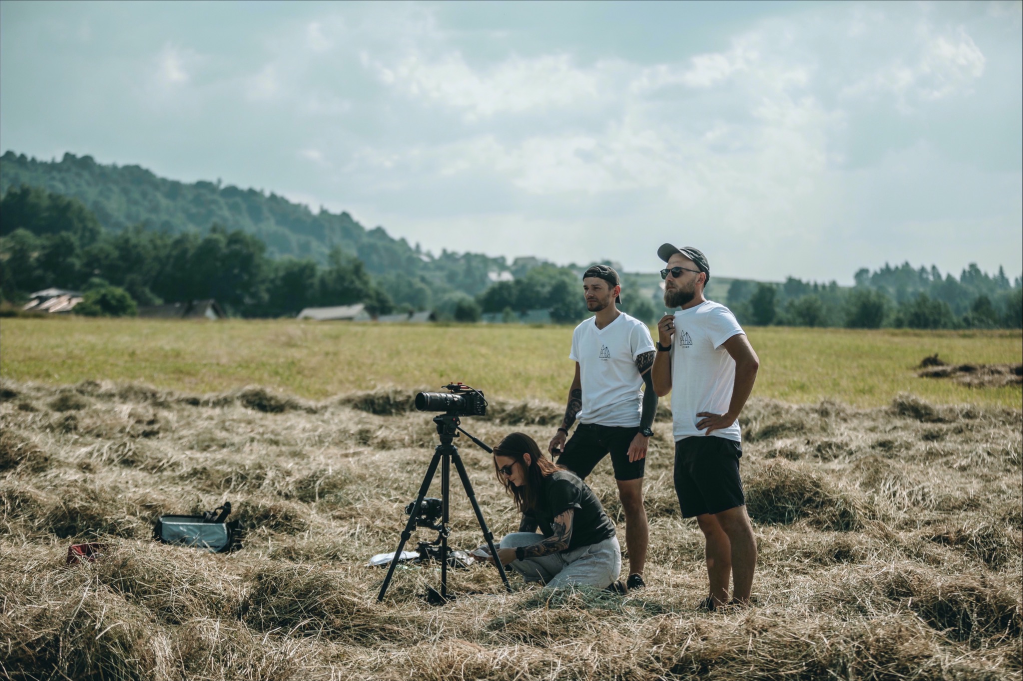 Ekipa filmowa: operator kamery na kolanach przy statywie z kamerą, dwóch mężczyzn w tle obserwuje, łąka z sianem i zalesione wzgórza w tle.