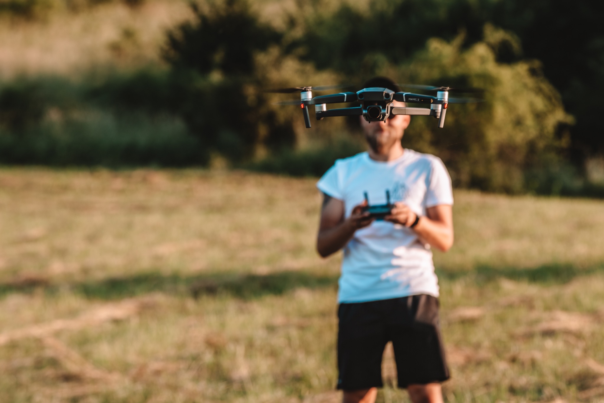 Operator drona z kontrolerem w rękach, dron Mavic 2 Pro zawieszony w powietrzu na tle łąki i drzew.