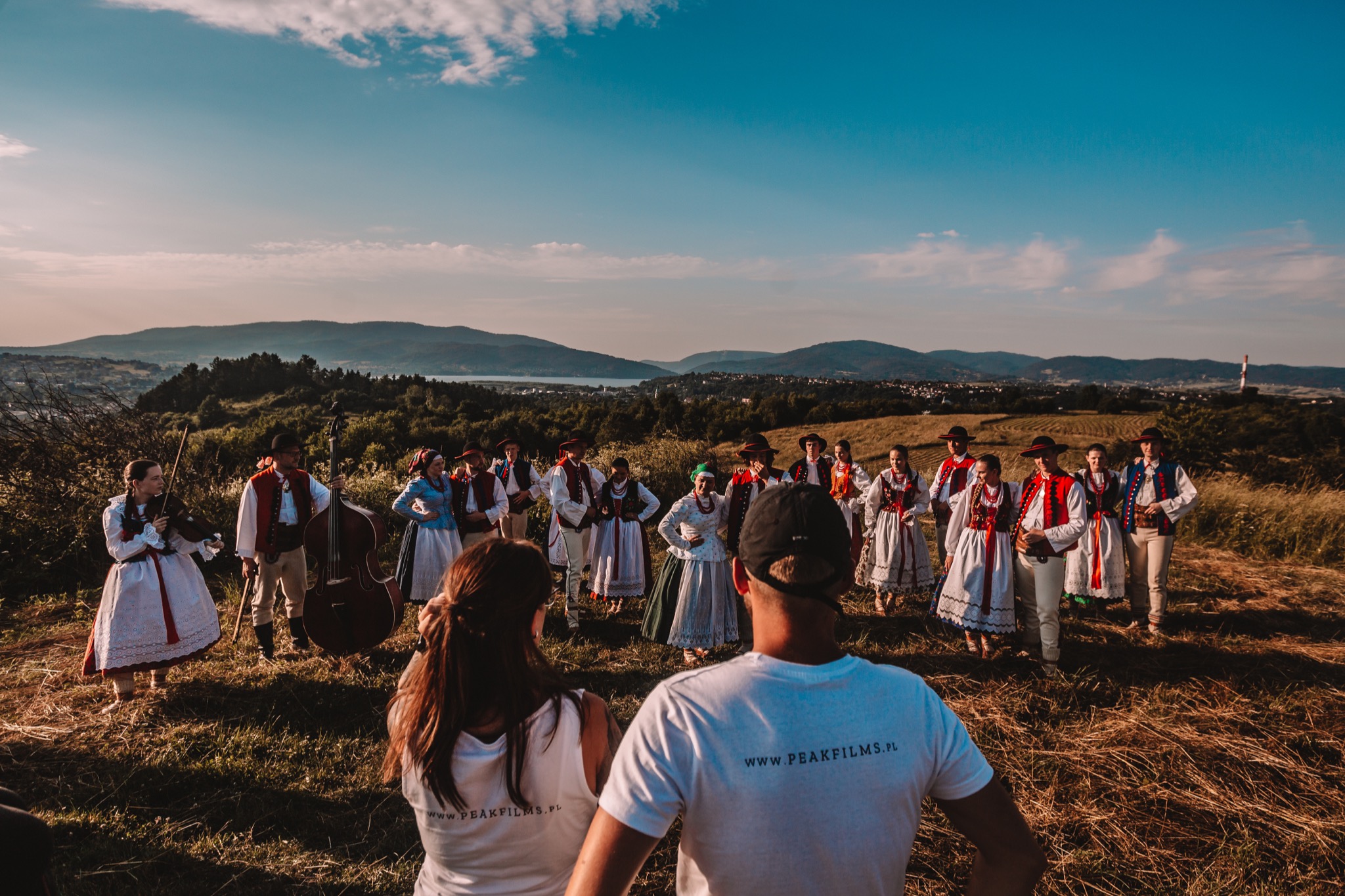 Ekipa filmowa Peak Films kręci ujęcia grupy w strojach ludowych na tle krajobrazu z górami i jeziorem.