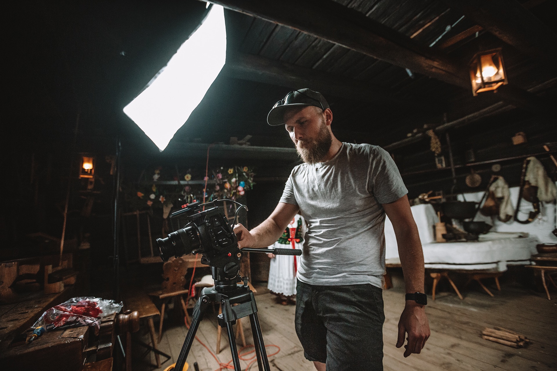 Operator kamery z brodą, w czapce, obsługuje profesjonalną kamerę na statywie w drewnianym wnętrzu, oświetlonym lampą, z elementami dekoracji ludowej w tle.