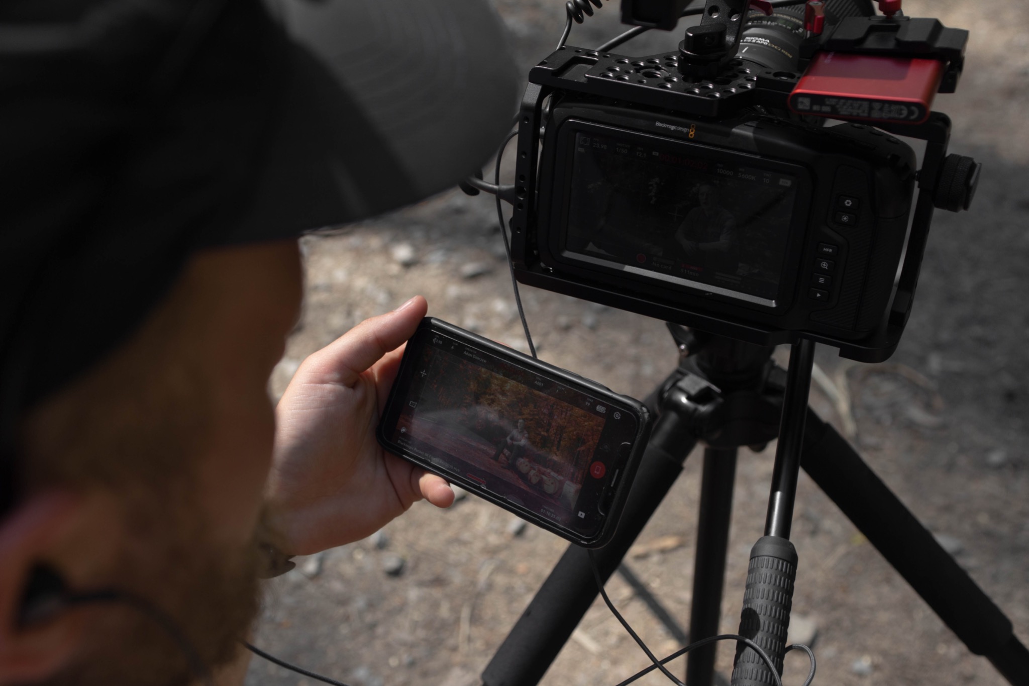 Operator sprawdza nagranie na smartfonie obok profesjonalnej kamery Blackmagic Design na statywie. Ekran kamery pokazuje podgląd nagrywanego materiału.