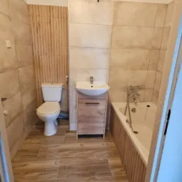 Ukończona łazienka z beżowymi płytkami imitującymi drewno na podłodze i ścianach, toaleta kompaktowa, umywalka nablatowa z szafką w kolorze jasnego drewna i wanna obudowana płytkami z pionowymi...