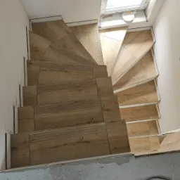 Spiralne schody wyłożone płytkami imitującymi drewno, widok z góry, z widocznym oknem dachowym i roboczym obuwiem na posadzce.