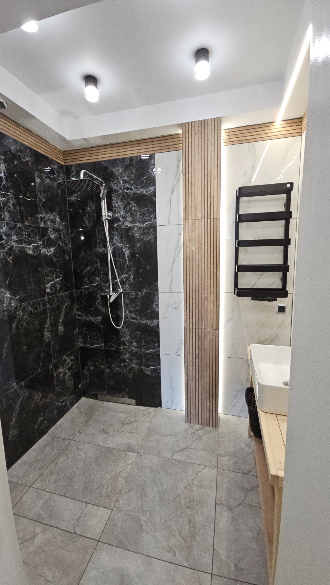 Nowoczesna łazienka z kabiną prysznicową wyłożoną czarnymi płytkami imitującymi marmur, szarymi płytkami podłogowymi i białą umywalką na drewnianym blacie.