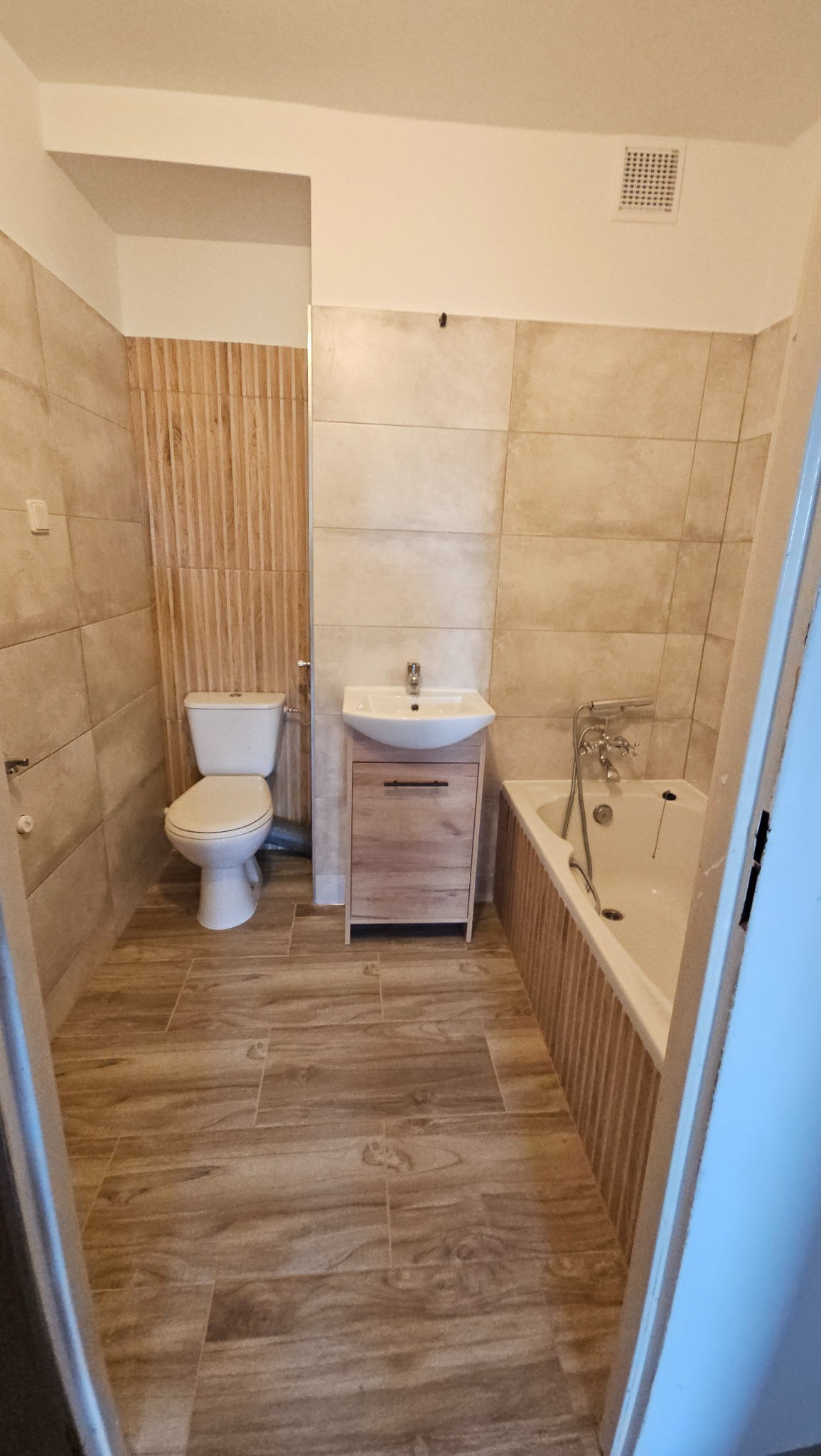 Ukończona łazienka z beżowymi płytkami imitującymi drewno na podłodze i ścianach, toaleta kompaktowa, umywalka nablatowa z szafką w kolorze jasnego drewna i wanna obudowana płytkami z pionowymi...