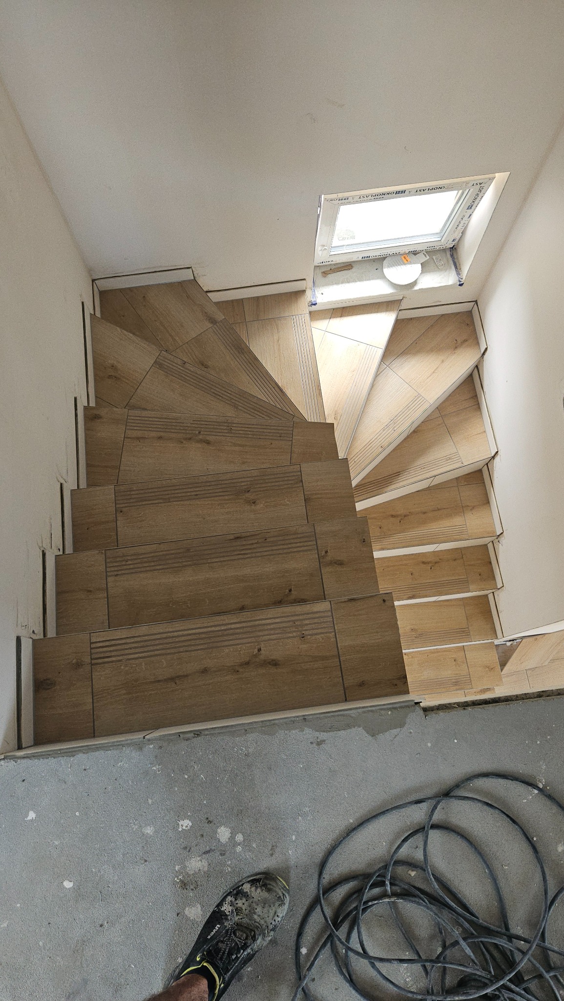 Spiralne schody wyłożone płytkami imitującymi drewno, widok z góry, z widocznym oknem dachowym i roboczym obuwiem na posadzce.