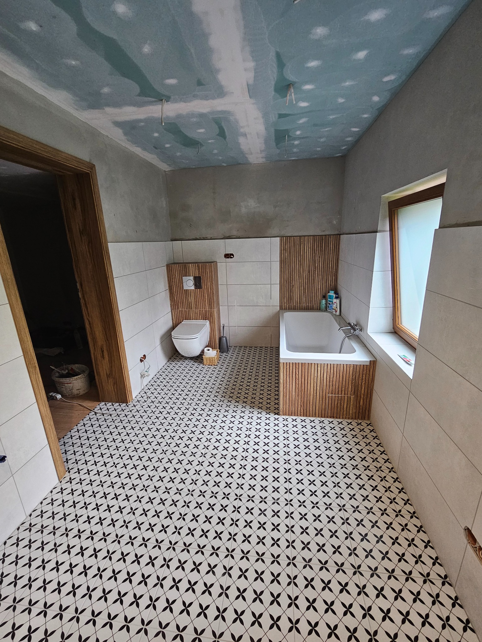 Łazienka w trakcie wykończenia: położone płytki podłogowe w czarno-biały wzór, częściowo wykończone ściany z białymi płytkami, obudowana drewnianymi panelami wanna i toaleta typu Geberit, sufit...