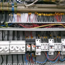 Rozdzielnica elektryczna w szarej obudowie z widocznymi bezpiecznikami, przewodami i złączkami oznaczonymi żółtymi numerami.