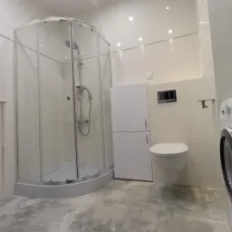 Nowoczesna łazienka z kabiną prysznicową narożną, białą zabudową meblową, podwieszaną toaletą i pralką, wykończona w jasnych odcieniach szarości i bieli.